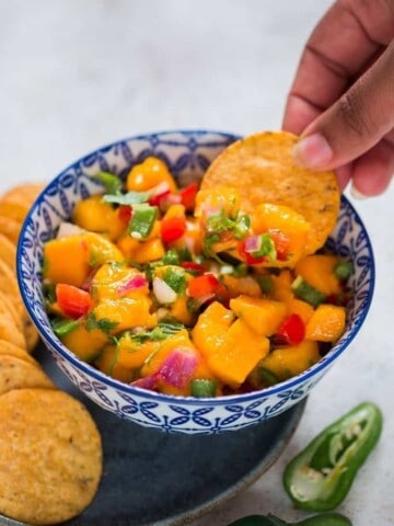 Finger scooping out mango salsa from a blue bowl with nachos chip