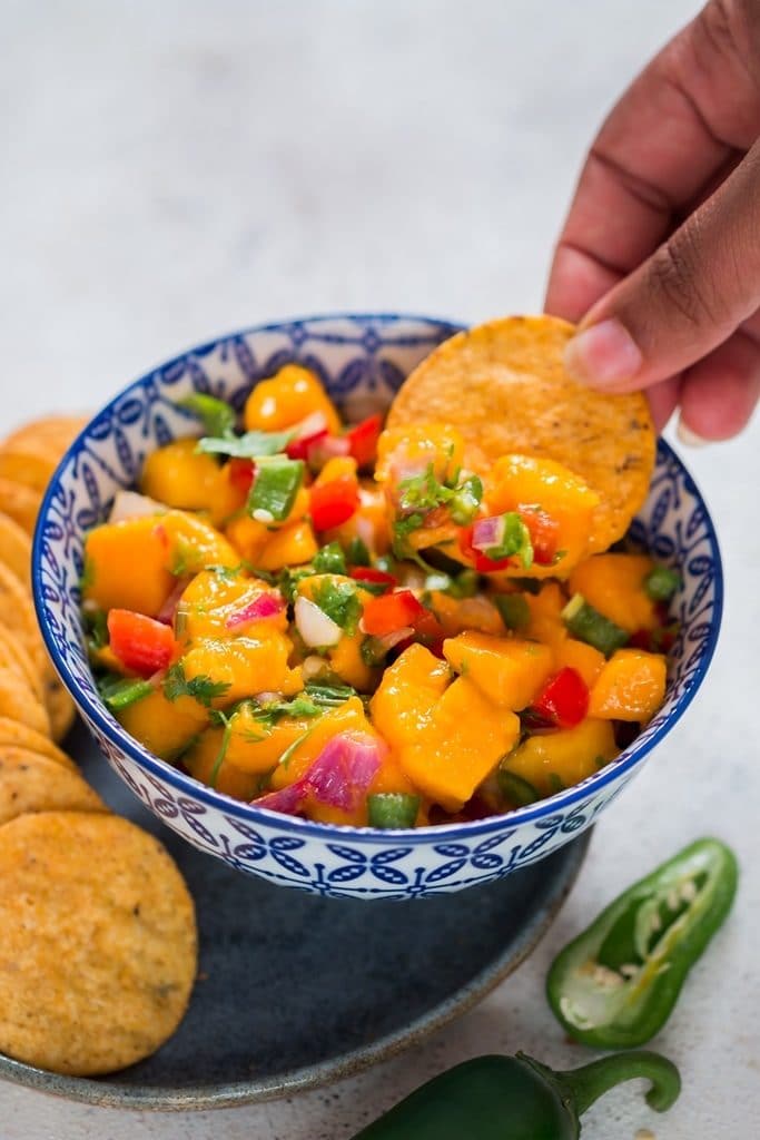 Finger scooping out mango salsa from a blue bowl with nachos chip