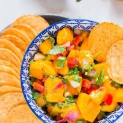 mango salsa recipe in blue stripped bowl with nacho chips arranged on a plate.