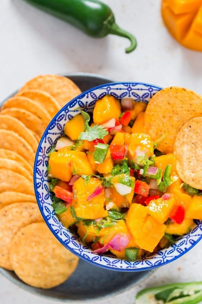 mango salsa recipe in blue stripped bowl with nacho chips arranged on a plate.