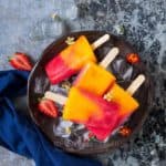 4 strawberry mango popsicles placed on round metal tray with ice cubes and pretty flowers on it