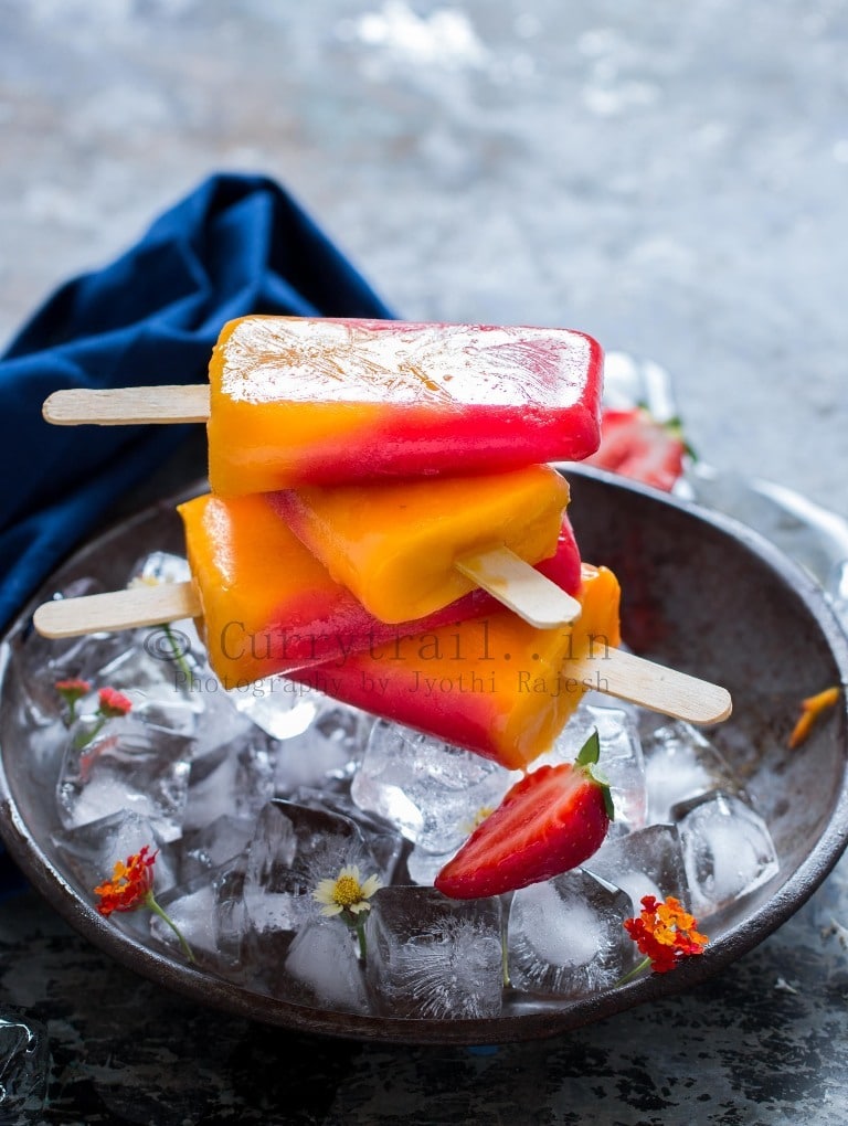 strawberry mango popsicles made of 2 ingredients stacked on top of each other on a plate full of ice cubes