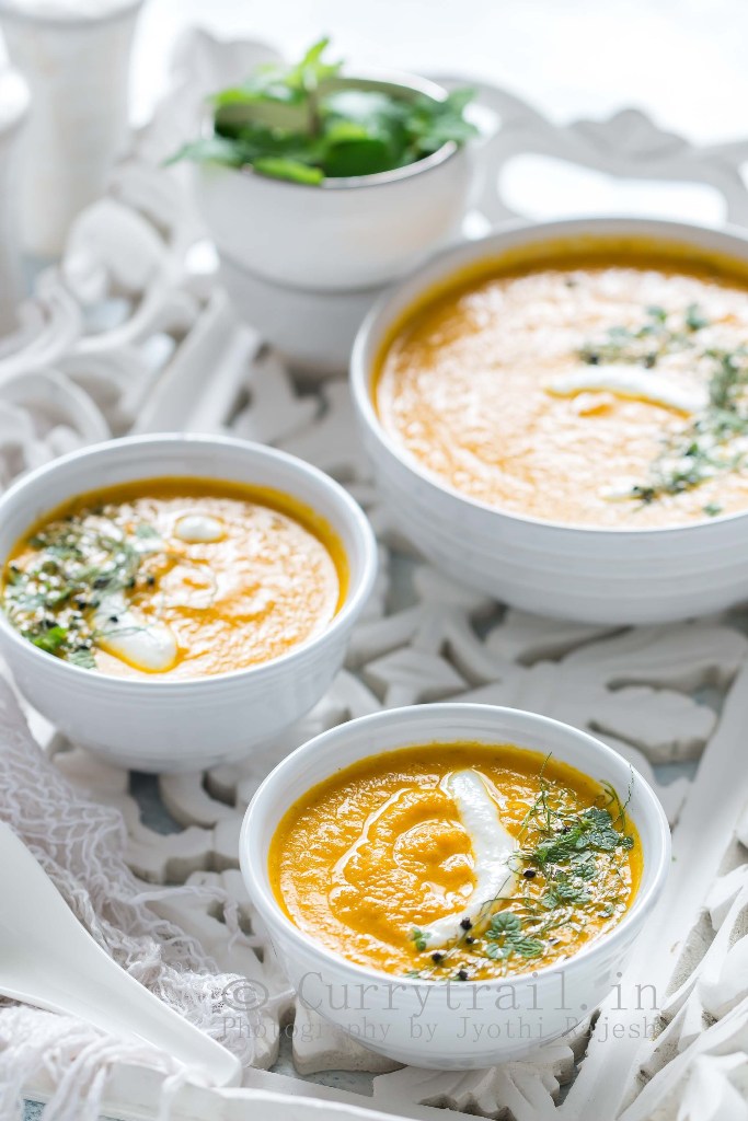 3 white bowls of roasted carrot soup served on white tray with roasted bread on side