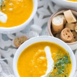3 white bowls of roasted carrot soup served on white tray with roasted bread on side