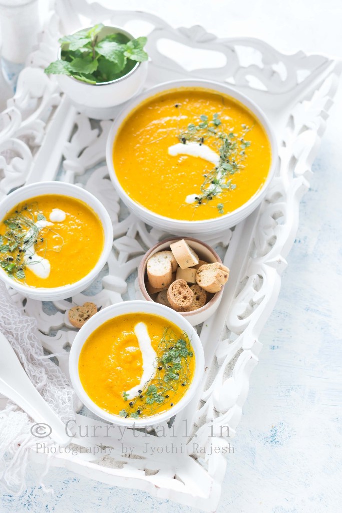 3 white bowls of roasted carrot soup served on white tray with roasted bread on side