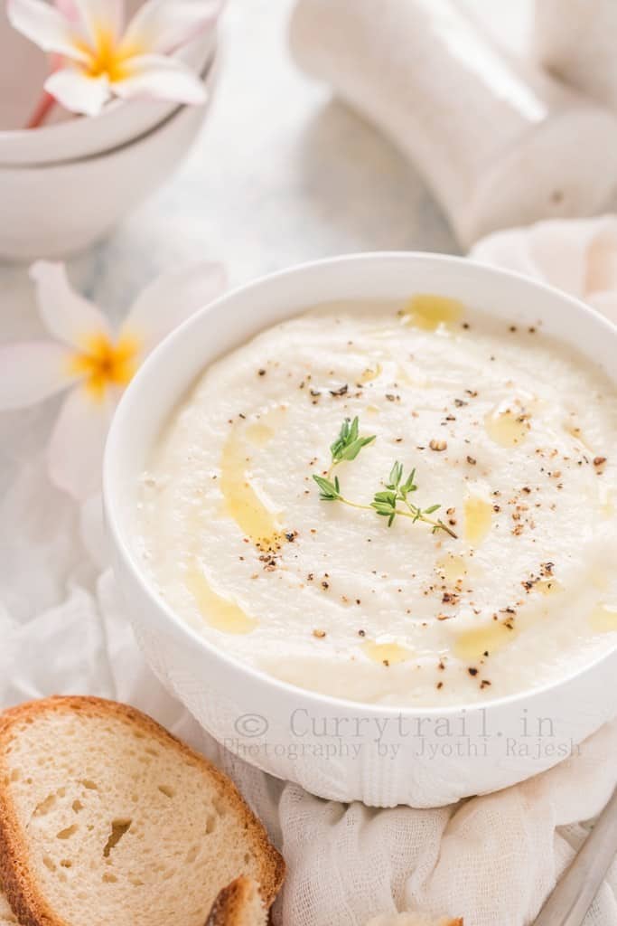 Cream of cauliflower soup in white bowl with crusty bread on sides and white petal flowers for the soul