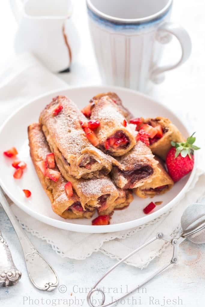 Nutella Strawberry French toast Roll Ups stacked up on white plate with morning coffee on the back