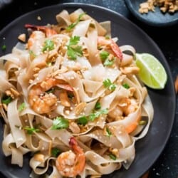 black plate with pad thai noodles garnished with peanuts, coriander and lime wedges on the sides