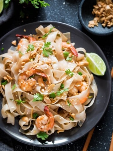 black plate with pad thai noodles garnished with peanuts, coriander and lime wedges on the sides