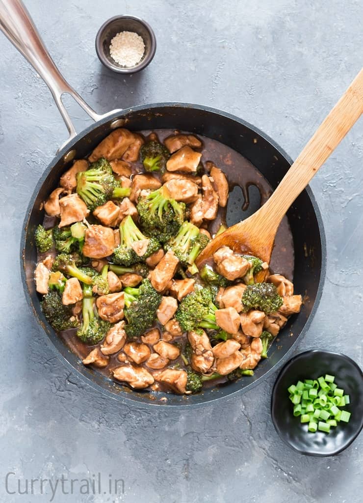 Chicken and broccoli stir fry in a skillet with wooden spoon and green onions sesame seeds on the sides