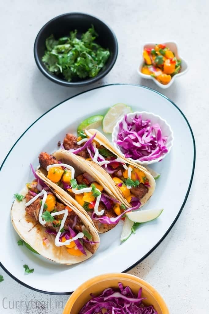 tilapia fish tacos lined in a white oval enamel plate with purple cabbage and mango salsa on small bowl in the side