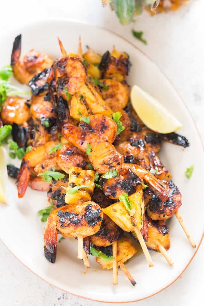 pineapple and shrimp skewered into bamboo sticks and grilled over BBQ placed on white oval plate with lemon wedges and cilantro leaves on top
