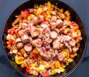 Sweet potato hash cooked in cast iron pan with browned sausages and bacon on top