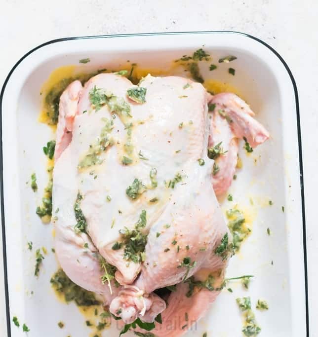 whole chicken rubbed with fresh herbs and butter with legs tied and placed on a white tray.
