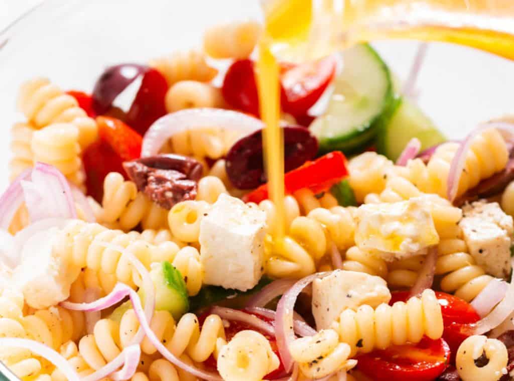 all ingredients for Greek pasta salad in a bowl with salad dressing poured over it.