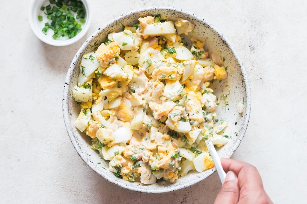 mixing in egg salad with its dressing in a white bowl.