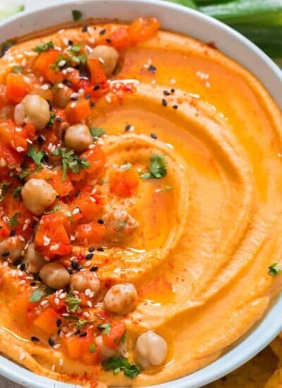 creamy roasted red pepper hummus made from scratch served in ceramic bowl with veggies and chips on sides
