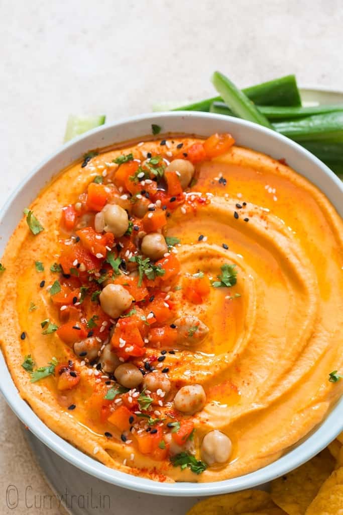 creamy roasted red pepper hummus made from scratch served in ceramic bowl with veggies and chips on sides