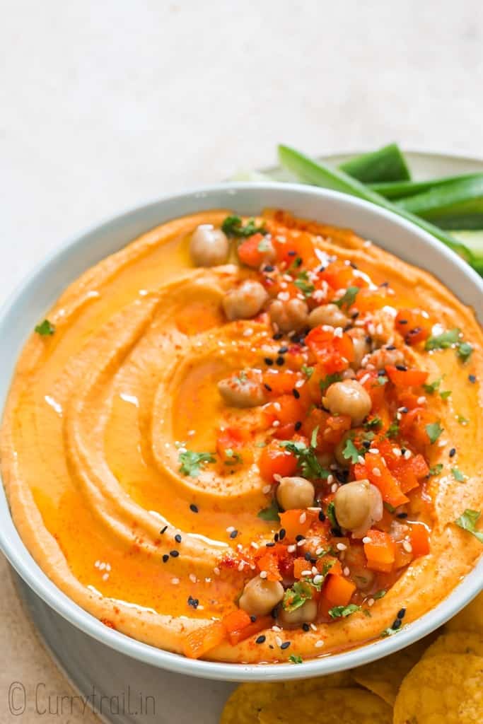 creamy roasted red pepper hummus made from scratch served in ceramic bowl with veggies and chips on sides