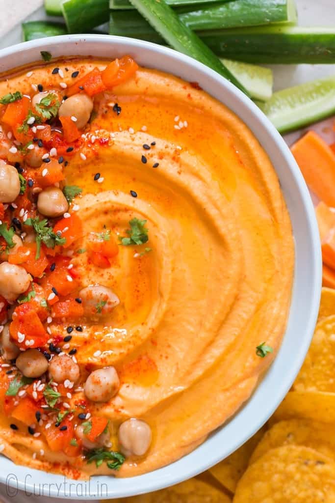 creamy roasted red pepper hummus made from scratch served in ceramic bowl with veggies and chips on sides