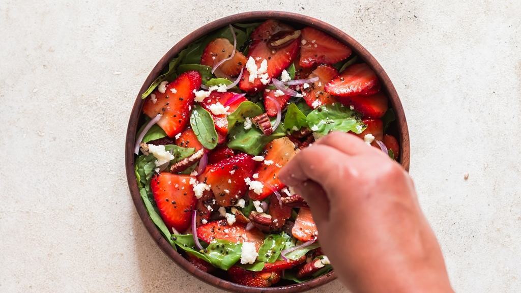 sprinkle feta cheese over strawberry salad in a wooden bowl.