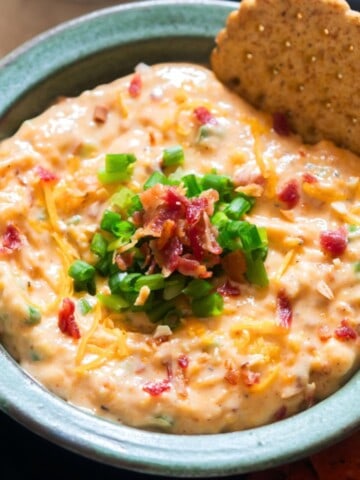million dollar dip in a bowl with green onion and crispy bacon on the top with cracker dipped in it.