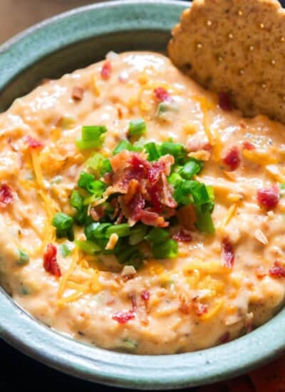 million dollar dip in a bowl with green onion and crispy bacon on the top with cracker dipped in it.